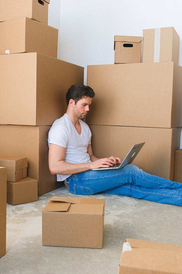 services-02 Young man using a laptop surrounded by stacked moving boxes indoors, depicting multitasking during relocation.